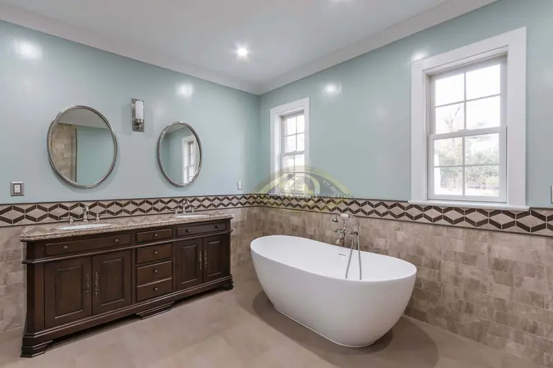 A bathroom with a dark wood double vanity under two round mirrors, a freestanding tub and stone wall tile with a geometric border