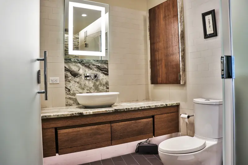 A bathroom with a floating walnut vanity, a vessel sink on a stone counter and a lit mirror