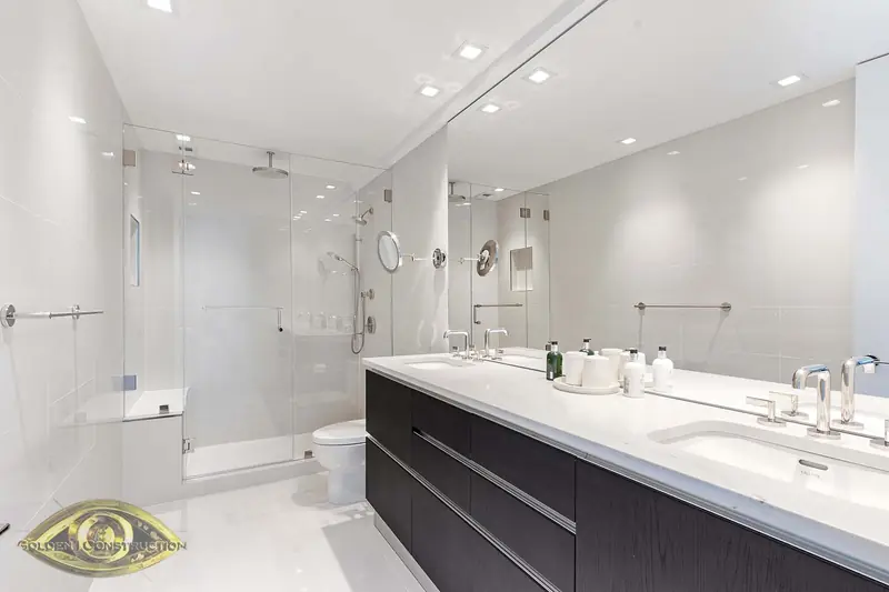 A white bathroom with a long dark double vanity under a white top, a wall-width mirror and a frameless glass shower