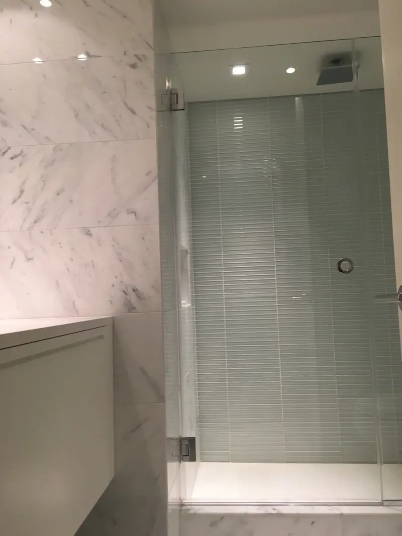 A shower with white marble walls, a frameless glass door and a back wall of green glass tile
