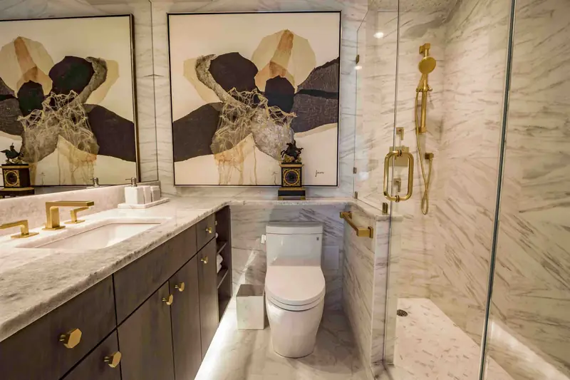 A marble bathroom with a dark vanity on brass knobs, a brass faucet, two abstract prints and a glass shower with brass fittings
