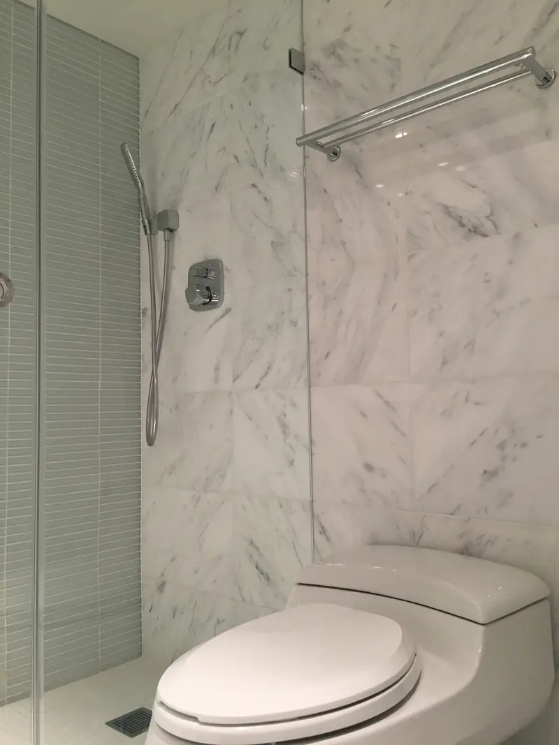 A marble-walled bathroom with a hand shower on a rail, a towel bar, a glass panel and a toilet