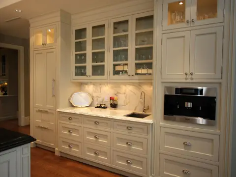 A wall of cream inset cabinets with lit glass-front uppers, a marble backsplash, a bar sink and a built-in coffee machine
