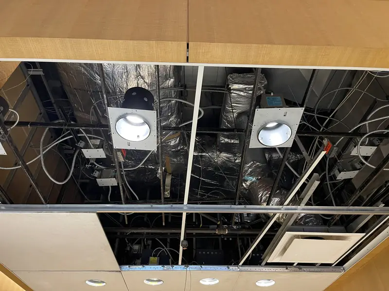 A ceiling opened up to show ducts, wiring and two recessed light housings on a black grid, above wood wall panels