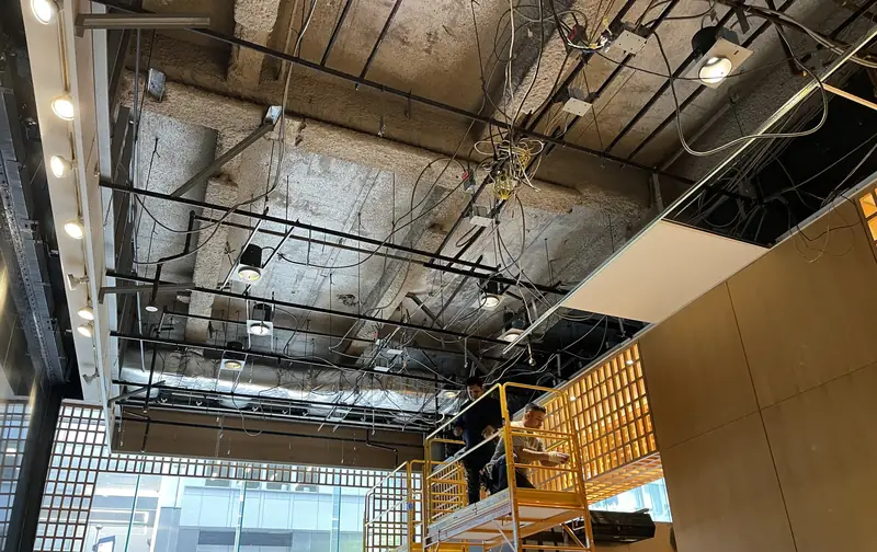 An entrance lobby with its ceiling opened to the ducts and wiring, a wood lattice wall, a scaffold and a glass front