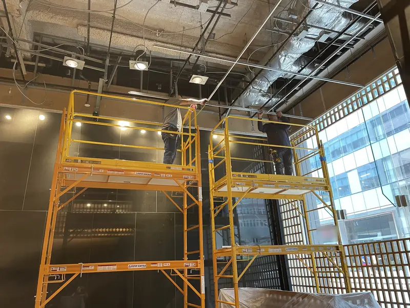 Two yellow rolling scaffolds under an open ceiling of exposed ducts and wiring, in a lobby with a wood lattice screen and a glass front