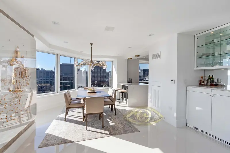 A white dining area with a table and chairs under a branching chandelier, corner windows onto the city and glass-front wall cabinets
