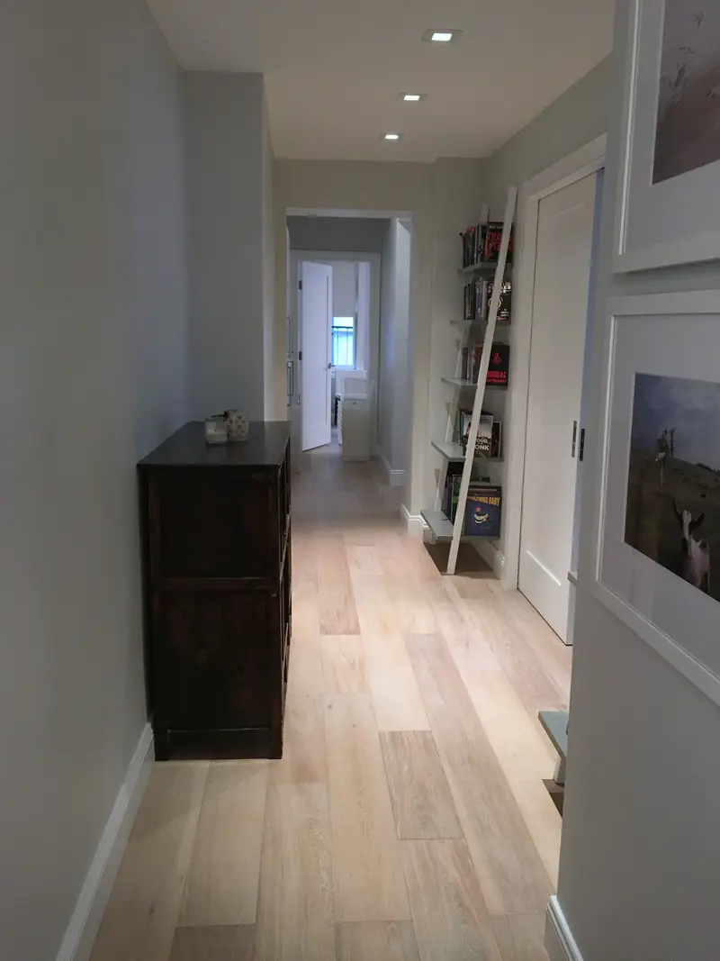 A long hallway with a pale oak floor, a dark chest and a leaning ladder shelf of books