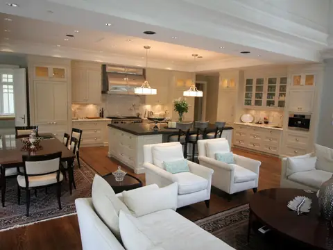 An open living, dining and kitchen room with cream cabinets to the ceiling, a dark island with stools and two pendant lights