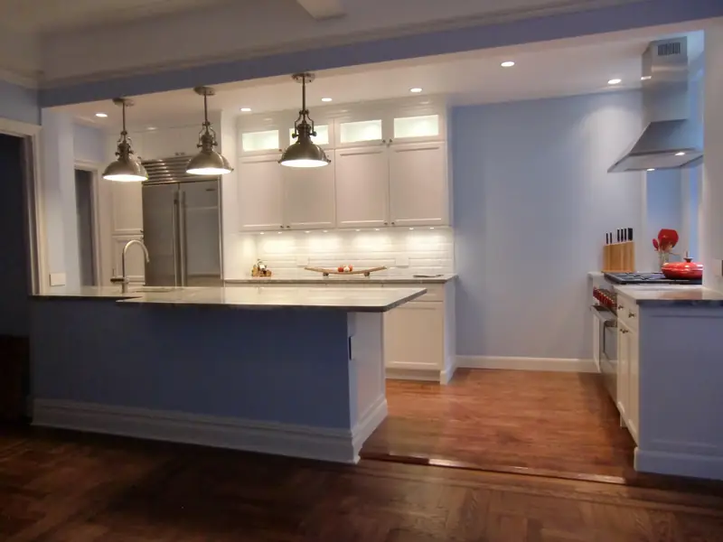 A kitchen with white cabinets and lit glass-front uppers, three metal pendants over a peninsula and pale blue walls