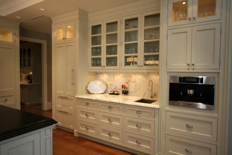 A wall of cream inset cabinets with lit glass-front uppers, a marble backsplash, a bar sink and a built-in coffee machine