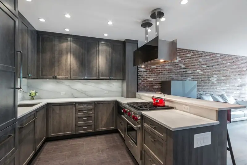 A U-shaped kitchen with dark grey stained shaker cabinets, white stone counters and backsplash, and an exposed brick wall