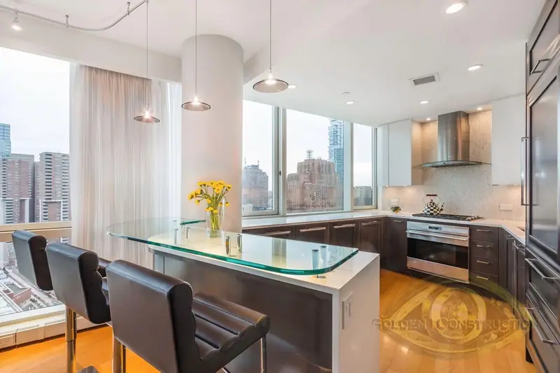 A high-rise kitchen with a glass breakfast counter and three black stools, dark cabinets, a round column and windows onto city towers