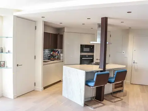 A white kitchen with a marble waterfall island, two blue stools, a walnut column and built-in ovens