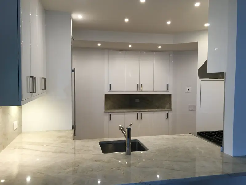 A polished stone counter with an undermount sink, facing white lacquer wall cabinets and a stone niche