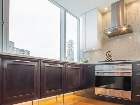 Dark wood base cabinets and a built-in oven under windows onto the city, with a steel and glass hood and a mosaic backsplash