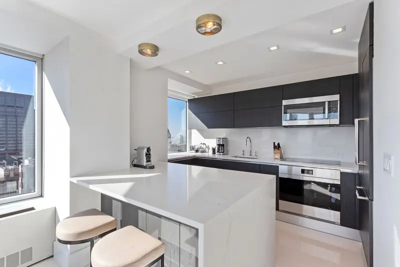 A white kitchen with a waterfall peninsula, dark wall cabinets, a built-in oven and microwave and windows onto city towers