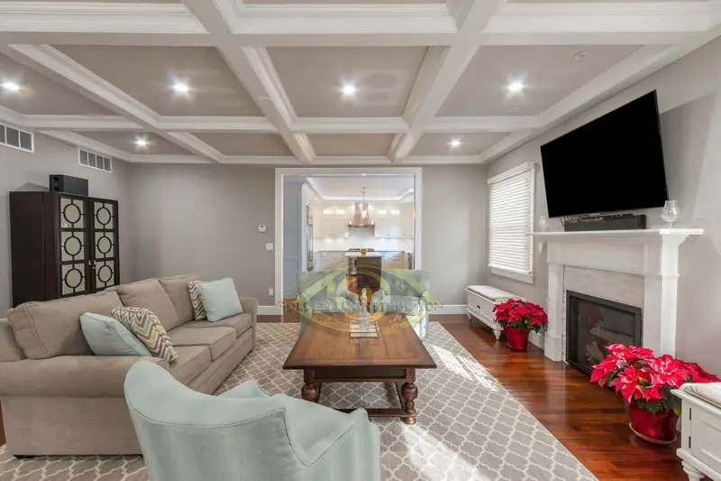 A living room with a white coffered ceiling, a television over a white fireplace mantel, a sectional sofa and a view through to a dining room