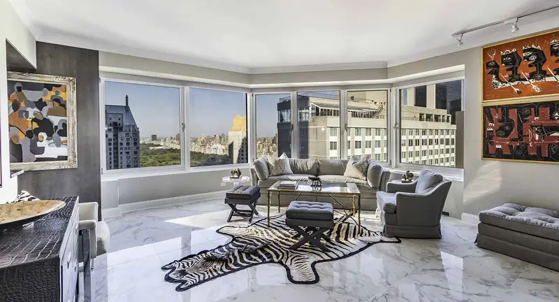 A living room with corner windows onto a park and city towers, grey sofas and a zebra hide rug on a white marble floor