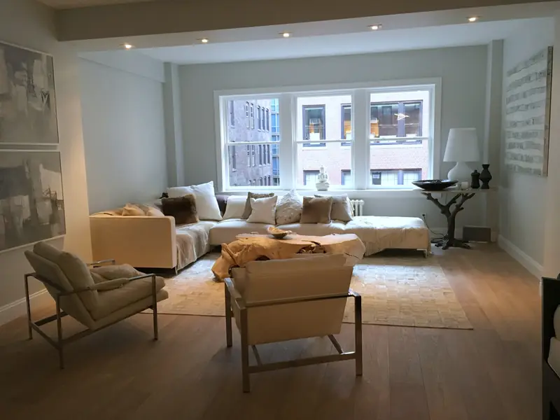 A living room with a pale sectional sofa, two lounge chairs and three windows, on a wood floor