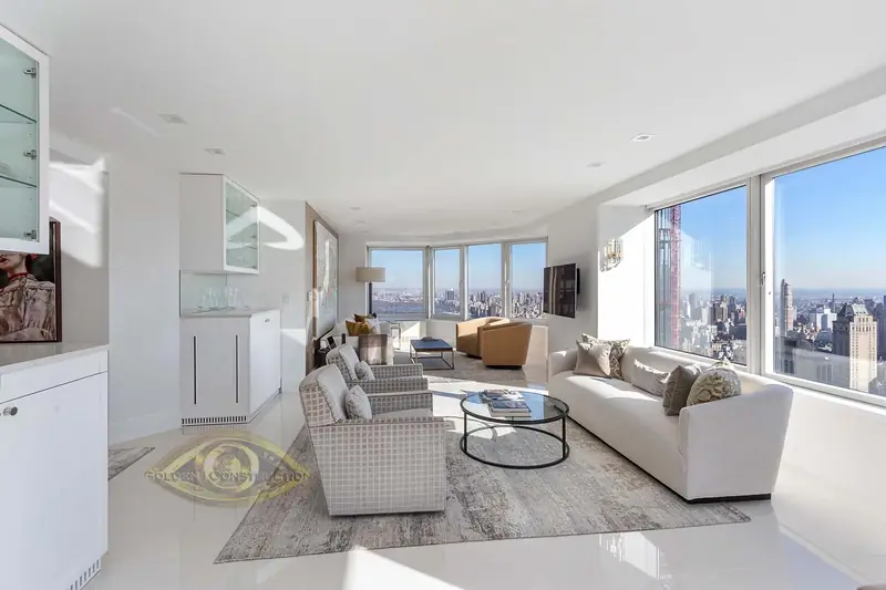 A white living room with a sofa and two armchairs around a round glass table, windows onto a skyline and white built-in cabinets