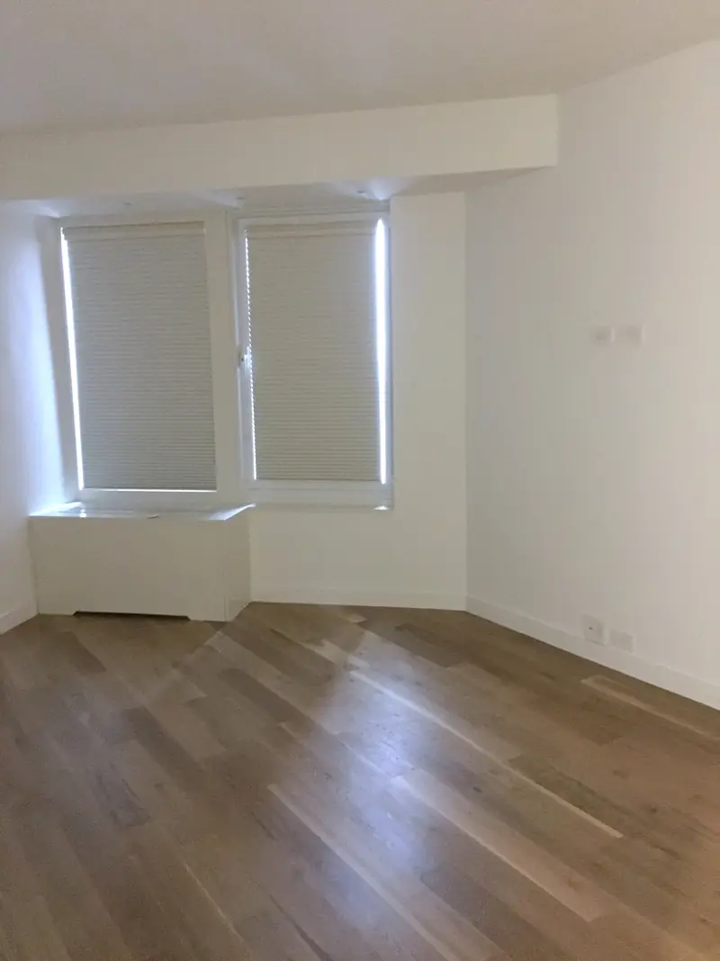 An empty room with an oak floor and two windows with shades over a low window seat