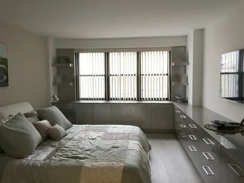 A bedroom with a run of built-in grey drawers under three windows with vertical blinds, and shelves at both ends