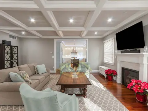 A living room with a white coffered ceiling, a television over a white fireplace mantel, a sectional sofa and a view through to a dining room